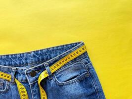 Close up of blue denim jeans with yellow measuring tape belt on vivid yellow background with copy space. Fashion, dieting, and body measurements. Top view. photo