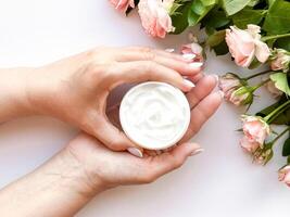 Hands middle age woman hold an open jar of white hand or body cream next to pink tea roses on white background, top view. Flat lay composition. Skincare and beauty product concept. photo