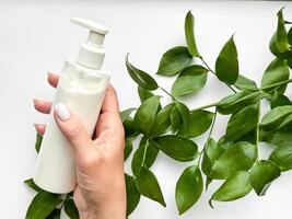 Close up of middle age woman hand holding white lotion bottle with green leaves on white background. Skincare and natural beauty concept. Healthcare, wellness, and organic product poster. photo