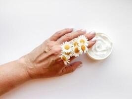 Hand middle age woman holding chamomile flowers next to an open jar of white hand cream on white background. Top view with copy space. Skincare, wellness and beauty product concept. photo