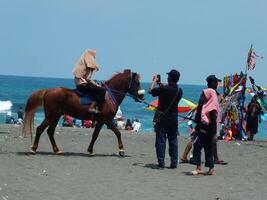 kebumen, central Java, Indonesia abril 11, 2024 personas disfrutar vacaciones montando un caballo durante brillante luz a ambal playa. verano familia turista Mancha con Oceano ola antecedentes. foto
