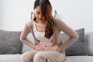 el mujer con un dolor de estómago es participación su estómago en dolor, sentado en el sofá en el vivo habitación. foto