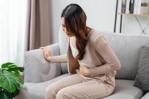 el mujer con un dolor de estómago es participación su estómago en dolor, sentado en el sofá en el vivo habitación. foto