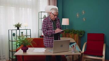 Senior grandfather man working on laptop computer making notes on sheet of paper calculating bills video