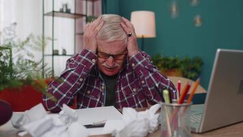 Senior man working on laptop computer, making notes on sheet of paper, upset by poor bad results video