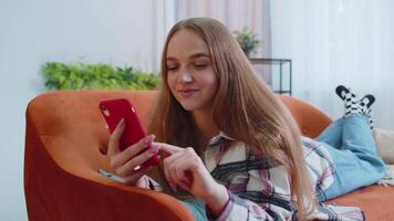 Joyful young adult woman lying on sofa using smartphone watching , online shopping, tapping video