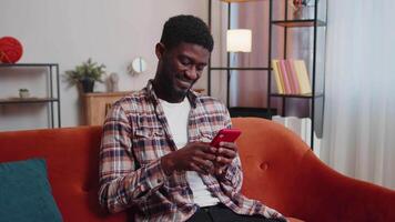 Joyful young adult man sitting on sofa using smartphone watching , online shopping, tapping video