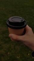 A hand holding a cup of coffee in their hand, with background of the green grass, mobile photo, vertical photo