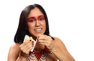 sonriente morena modelo en brasileño indio disfraz y maquillaje participación un tapioca, un típico brasileño bocadillo foto