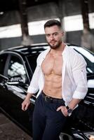 Handsome young sexy man near black car. Macho man in unbuttoned shirt near luxury auto. Selective focus. Closeup. photo