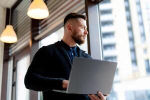 Cute young businesman works on hi tech lap top in a cafe. Male looks at window with personal computer in hands. photo