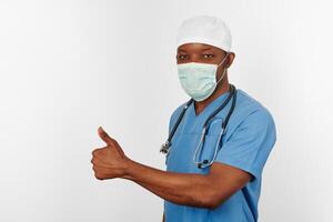 Black surgeon doctor man in blue coat white cap and surgeon mask with thumbs up gesture photo