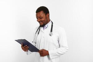 Smiling black bearded doctor man in white robe with stethoscope looks to medical chart on clipboard photo