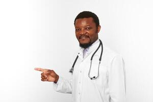 Smiling black bearded doctor man in white coat with stethoscope points finger, white background photo