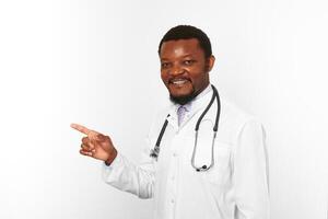 Smiling black bearded doctor man in white coat with stethoscope points finger, white background photo