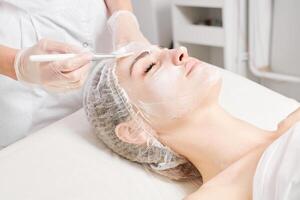 Beautician applies vitamins cream mask on woman face for rehydrate face skin in beauty salon photo