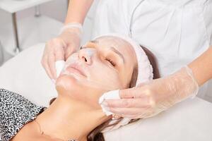cosmetólogo quitando facial crema máscara desde mujer cara piel con cosmético toallitas en belleza salón foto