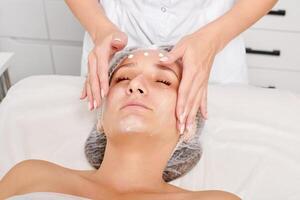 Cosmetologist rubbing moisturizing cream mask into woman face skin for rejuvenation in beauty salon photo