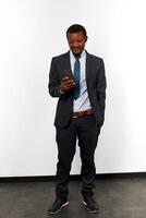 Smiling african american black man in business suit looks in smartphone white wall background photo