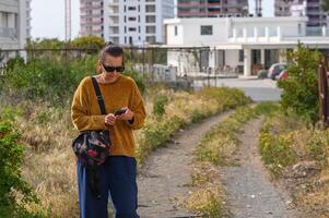 woman holding a mobile phone with, standing or walking against the background of houses. 1 photo