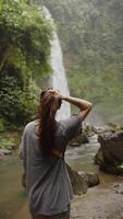 fille permanent retour à le caméra à la recherche à une cascade dans le jungle, verticale video