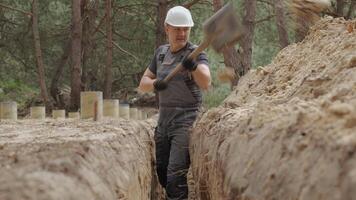 A worker wearing a white hard hat is actively digging a narrow trench in a forested area. The surrounding terrain is uneven with visible tree stumps and soil heaps. video