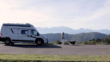 Man in jacket playing with his dog nearby a white van against mountains, contemplate thoughts and find calm peace. Camping trip in great outdoors. Van lifestyle. Family vacation travel video