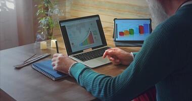 Home Office. A man with glasses looks at graphs on a laptop and tablet and writes notes in a blue notebook video