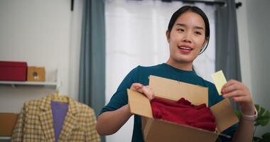 Aufnahmen Dolly Schuss, jung asiatisch Frau Geschäft Inhaber Stand Verkauf Kleider Leben Streaming auf Handy, Mobiltelefon Telefon und verpackt Kleider in Box zum ein Kunde ,online Einkaufen video