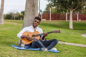 un africano americano hombre se sienta debajo un árbol y canta en naturaleza mientras jugando un guitarra. foto