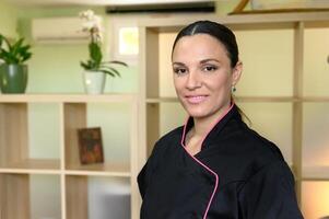 portrait of massage therapist smiling looking at camera, standing in professional space such as a spa, salon, or massage room. shelving unit with various compartments on background. photo