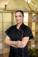 massage therapist looking at camera, standing with her arms crossed in professional space such as a spa, salon, or massage room. shelving unit with various compartments on background. photo