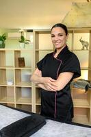 confident massage therapist wearing black top smiling looking at camera standing with her arms crossed next to massage counter in massage room. Place beauty or health-related services are provided. photo