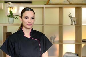 portrait of massage therapist looking at camera, standing in professional space such as a spa, salon, or massage room. shelving unit with various compartments on background. photo