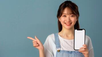 Cheerful woman showing smartphone. An engaging young Asian woman in overalls, smiling and pointing at a blank smartphone screen, ideal for app promotion. . photo