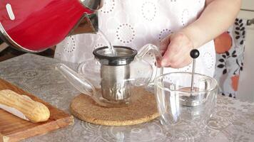 Woman pours hot water from electric red kettle into glass teapot. On table vanilla eclair cake, cup. video