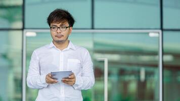 A man wearing a white shirt and glasses is holding a tablet in his hand photo