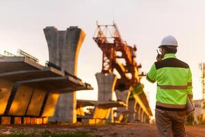 A construction worker is standing on a bridge, talking on his cell phone. The scene is set in a construction site, with a large crane in the background. The worker is wearing a yellow jacket photo