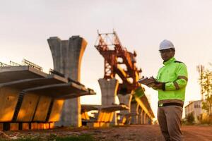 A construction worker is standing on a bridge, talking on his cell phone. The scene is set in a construction site, with a large crane in the background. The worker is wearing a yellow jacket photo