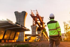 A construction worker is standing on a bridge, talking on his cell phone. The scene is set in a construction site, with a large crane in the background. The worker is wearing a yellow jacket photo