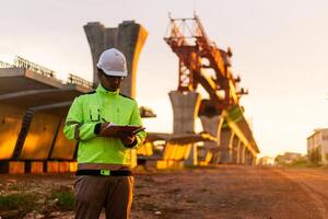 A construction worker is standing on a bridge, talking on his cell phone. The scene is set in a construction site, with a large crane in the background. The worker is wearing a yellow jacket photo