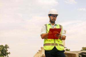 un hombre en un amarillo chaqueta es escritura en un portapapeles mientras en pie en frente de un puente. concepto de trabajo y productividad foto