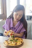 un mujer es comiendo un Pizza con su manos. el Pizza es en un de madera lámina. el mujer es vistiendo un púrpura camisa foto