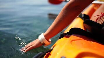 A woman is sitting in a kayak on a lake. The kayak is orange and the paddle is red. The woman is wearing a bikini and she is enjoying her time on the water. video