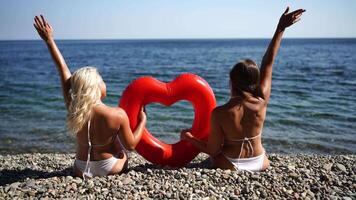 Two women sitting on a beach holding a red heart shaped inflatable pool toy. Scene is lighthearted and fun. video