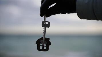 Keys with a keychain in the shape of a house in a hand in black gloves on the background of the sea. The concept of buying a house, apartment, real estate, young family, mortgage loan, rental housing. video