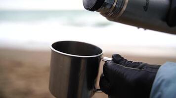Unrecognizable woman pours water into a mug from a thermos near the sea, Slow Motion, closeup on a cup and thermos. The woman is wearing black gloves and a blue jacket. Cinematic frame. video