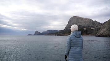 un mujer turista levanta su manos arriba, en pie en un muelle terminado el mar en contra el antecedentes de el cielo y montañas, vestido en un azul chaqueta. observación de el hermosa paisaje en el costa. video