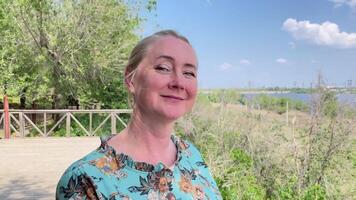 Middle aged blonde woman with long braided hair looks at the camera and smiles against the background of a park by the river on a summer day, slow motion, video