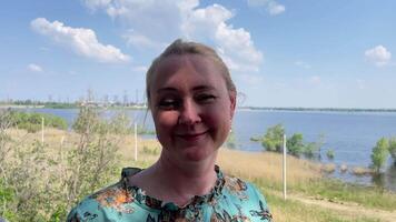 Middle aged blonde woman with long braided hair looks at the camera and smiles against the background of a park by the river on a summer day, video